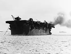 Photograph of badly damaged aircraft carrier, afloat with smoke coming from deck.