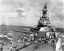 A large ship tilted to the right, with gun barrels pointed to the left: Crewmen can be seen on the battleship's deck. To the left of the image another large warship can be seen.