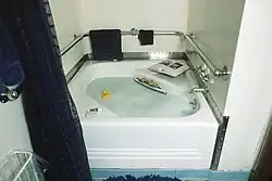 A small bathtub is visible in the center of the image. Bubbles, a rubber ducky, and a small floating boat can be seen in the tub, while two books, a soap bar, and a toothpaste tube can be seen around the rim of the bathtub.