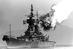A black and white image; a large ship with gun barrels pointed to the right and up, flames and smoke can be seen emanating from the gun barrels