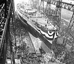 A large warship, still missing most of its superstructure, sits in a dry dock, awaiting its launch. The ship is draped in a large banner and surrounded by crowds of spectators; a huge gantry towers over the ship.