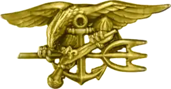 A gold image depicting an eagle perched on an anchor, clutching a trident with one claw and a gun in the other.