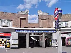 A brown-bricked building with a rectangular, light blue sign reading "METROPOLITAN & PICCADILLY LINES" in white letters all under a blue sky