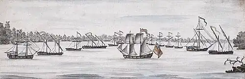 A group of 14 small ships of varying sizes on a lake with wooded land in the background