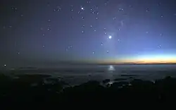 A photograph of the night sky taken from the seashore. A glimmer of sunlight is on the horizon. There are many stars visible. Venus is at the centre, much brighter than any of the stars, and its light can be seen reflected in the ocean.