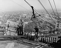 Two men are standing high in the air on a walkway, and a wheel is above them, suspended by wires.