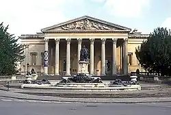 A Palladian style nineteenth century stone building with a large colonnaded porch. In front a large metal statue on a pedestal and fountains with decorations.