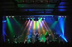 Members of a band, long-haired and dressed in black, perform under multi-colored lights.