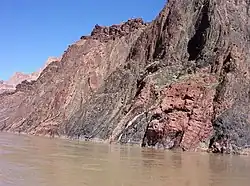 Gray and reddish rock face with rough surface adjacent to a river.