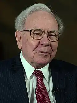 A chest level portrait of Warren Buffet against a dark background wearing a dark suit, a white shirt, and a red tie. He is unsmiling, looking past the camera.