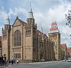 A grand gothic church-like building as viewed from a short distance away across a pedestrianised street. The building has a grand stained glass window at the front surrounded by two cuboid towers, and a large cuboid tower at the back-right with a red brick roof.
