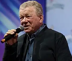 Headshot of William Shatner in GalaxyCon Richmond. He is a later-aged, clean-shaven, light-skinned man with grey hair. He is wearing a button-up shirt and a jacket on top.