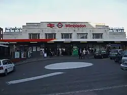 A white-bricked building with a sign reading "Wimbledon" in red letters and people walking in front all under a light blue sky with white clouds