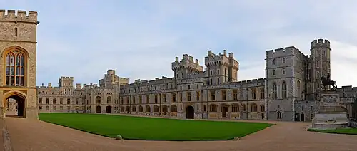 A photograph of a grey Gothic quadrangle with a green grass square in the middle. On the left, a block of the building makes up the near ground. A gatehouse is in the middle of the right-hand part of the quadrangle.