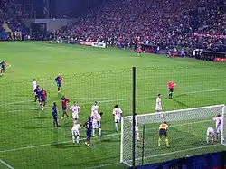 A photograph from behind the goal shows Barcelona and Wisła Kraków players gathering in the near penalty area for a corner kick. Wisła Kraków players are standing closer to the goal, and the referee is standing on the edge of the penalty area.