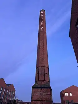 A photograph looking up from ground level at a tall, brick-built octagonal chimney on a square base with 'Wolsey' written vertically in white caps at the top.