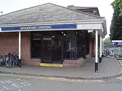 A red-bricked building with a rectangular, dark blue sign reading "WOODFORD STATION" in white letters all under a clear, white sky