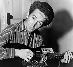Black-and-white photo of Guthrie looking up at an angle away from the camera and playing a guitar that says "This machine kills fascists"