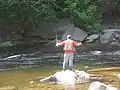A man in an orange shirt and tan vest fishes in a stream with rapids and large boulders