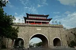 A wide, grey brick city wall featuring two large arched gateways for traffic. A colorful, multi-story pavilion (gate tower) with a red facade and dark tiled roof is mounted on top of the wall against a bright blue, cloudy sky.