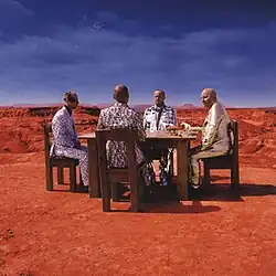 Four men wearing uniquely designed suits sitting by a table at Bardenas Reales with horses on the table and the Earth and Moon in the background.