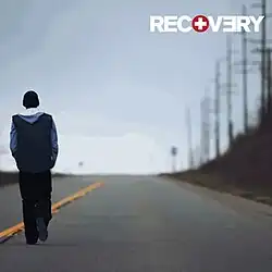 The cover features Eminem walking down the road under clear albeit cold looking blue sky. On top-right corner, in bold and capitalised format, the title RECOVERY appears.