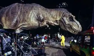 A life-sized Tyrannosaurus robotic model, with hydraulics where the dinosaur's feet would be, touches a car in a movie set.